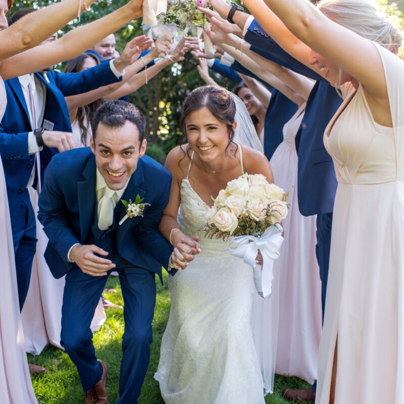 Bride and groom smiling under a floral arch made by friends.