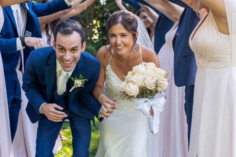 Bride and groom smiling under a floral arch made by friends.