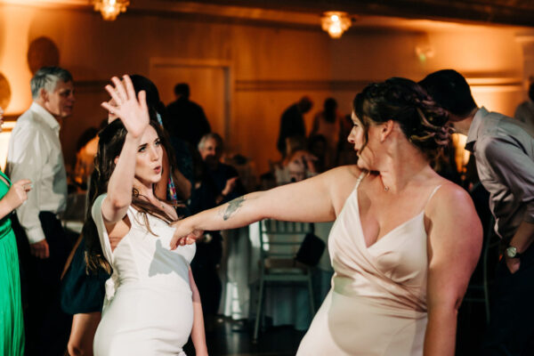 Two women dancing in a room with people standing around.