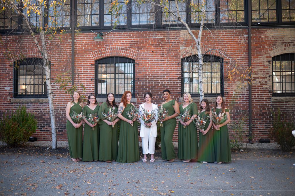Bridal party posing outdoors in front of a brick wall, bridesmaids in green dresses and groom in white suit. Bridal party posing outdoors in front of a brick wall, bridesmaids in green dresses and groom in white suit.