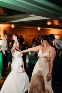 Two women dancing in a room with people standing around.