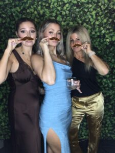 Three women holding fake mustaches in their mouths.