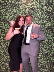 A man and woman posing for a picture in front of a wall covered with green leaves.