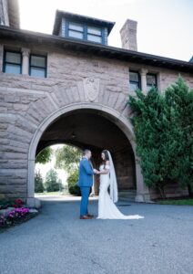 A couple in wedding attire stands under a stone archway outdoors.