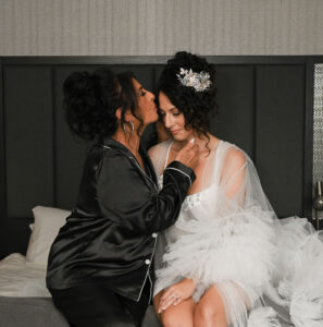 Two women sharing a tender moment in elegant attire, one in black satin and the other in a white bridal robe.