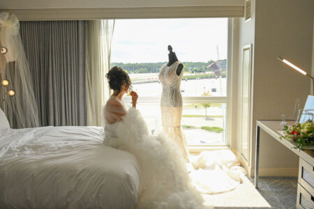Bride in a wedding dress sitting on a bed by a balcony with another dress hanging.