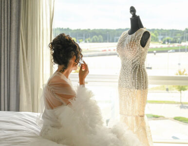 Bride in a fluffy robe looking at a wedding dress on a mannequin by a window.