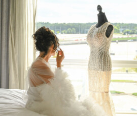 Bride in a fluffy robe looking at a wedding dress on a mannequin by a window.