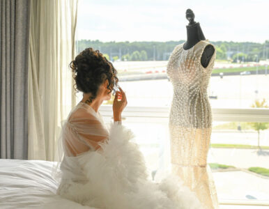 Bride in a fluffy robe looking at a wedding dress on a mannequin by a window.