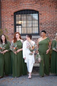 A bridal party with bridesmaids in green dresses and a bride in a white suit holding flowers.