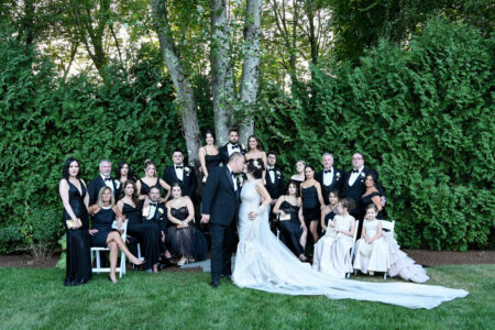 A formal wedding group photo with the bride and groom surrounded by guests in elegant attire outdoors.