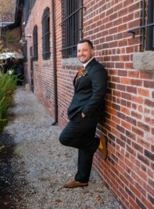 Man in a suit leaning against a brick wall, smiling.