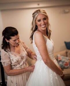 Bride smiling as a woman helps with her wedding dress.