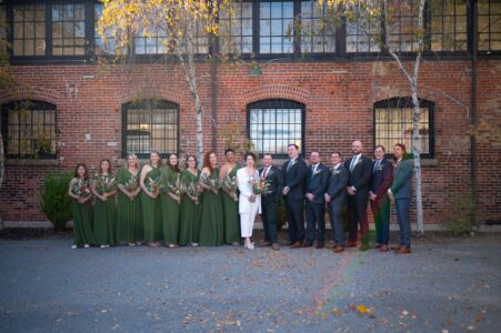 Group photo of wedding party with bridesmaids in green and groomsmen in suits outside a brick building.
