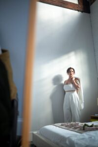 Woman in a white dress standing by a sunlit wall, looking thoughtful.