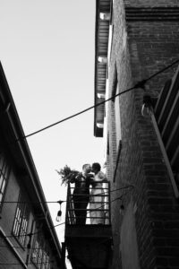 Couple embracing on a balcony between brick buildings, black and white.