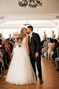 Newlyweds share a kiss at their wedding ceremony surrounded by guests.