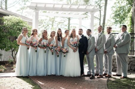 Bride and groom with bridal party in light attire under pergola.