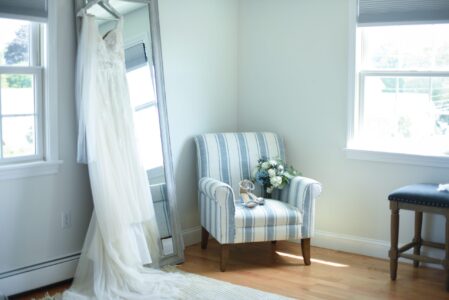 A wedding dress hangs beside a striped armchair with flowers in a bright room.
