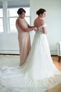A woman in a pink dress helps a bride with her wedding gown in a bright room.