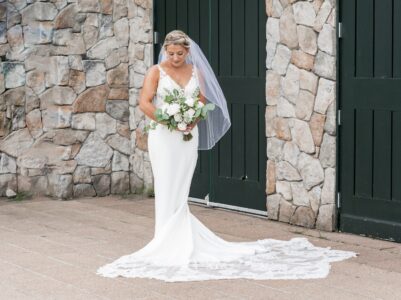 Bride in elegant white gown holding bouquet, standing before green doors and stone wall.