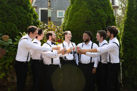Groom and groomsmen in white shirts and suspenders toasting with drinks outdoors.