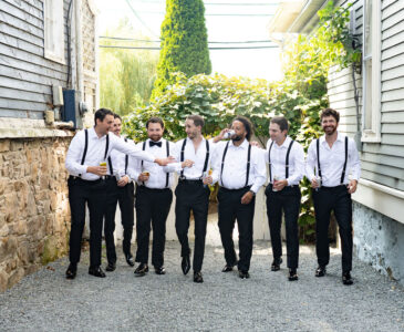 Seven men in formal attire with suspenders walking outdoors on a sunny day.