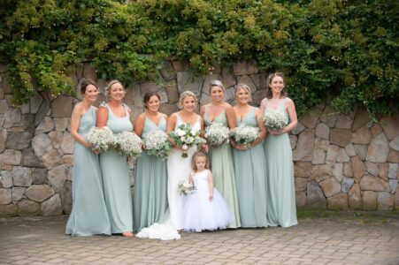 Bridesmaids in sage dresses with a flower girl in white, posing outdoors.