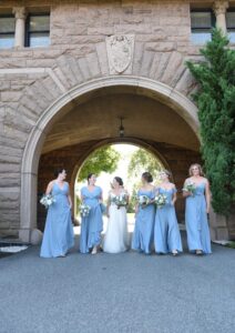 Bride and bridesmaids in blue dresses posing under stone archway.