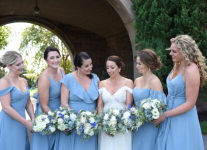 Bride with bridesmaids in blue dresses holding white and blue bouquets outdoors.