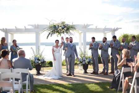 Bride and groom with wedding party under a floral arch outdoors.