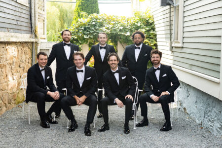 Seven men in black tuxedos posing outdoors with greenery behind.