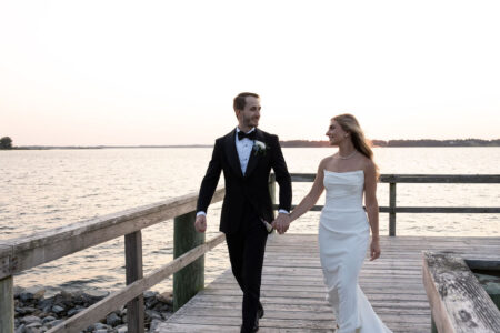 Newlywed couple holding hands, walking on a pier at sunset.