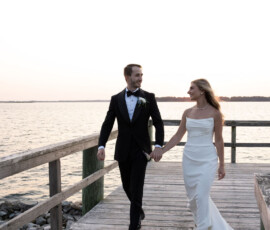 Newlywed couple holding hands, walking on a pier at sunset.